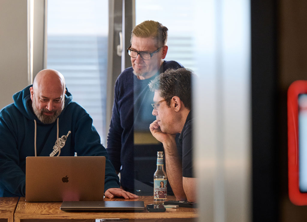 Drei Männer stehen über ein Macbook geneigt und programmieren gemeinsam.