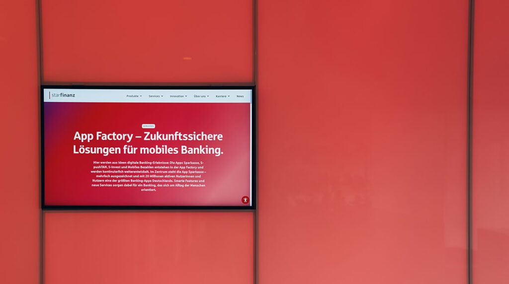 Ein Display vor einer roten Wand zeigt die App Factory Webseite