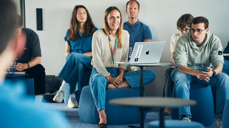 Eine Frau sitzt vor dem aufgeklappten Laptop und hält ein Meet Up und ihre Mitarbeitenden sitzen um sie herum und hören zu.