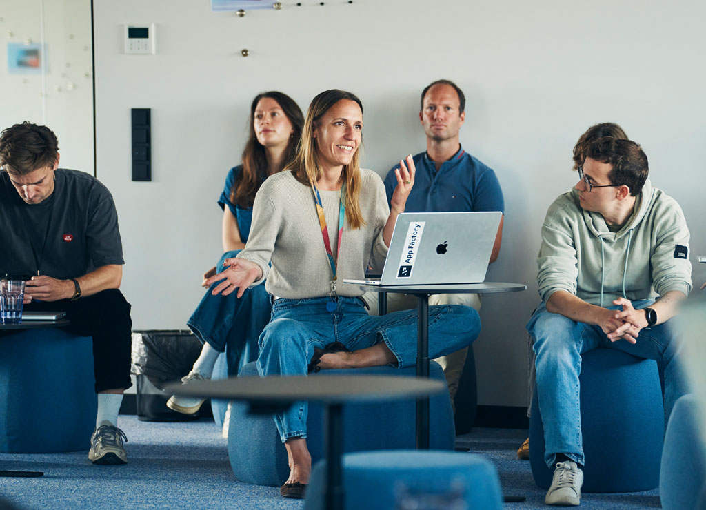 Eine Frau sitzt vor dem aufgeklappten Laptop und hält ein Meet Up und ihre Mitarbeitenden sitzen um sie herum und hören zu.