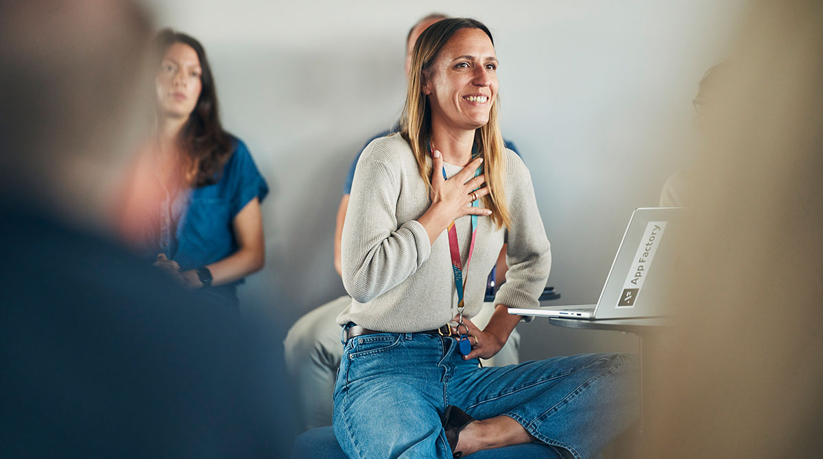 Eine Frau sitzt vor dem aufgeklappten Laptop und hält ein Meet Up und ihre Mitarbeitenden sitzen um sie herum und hören zu.