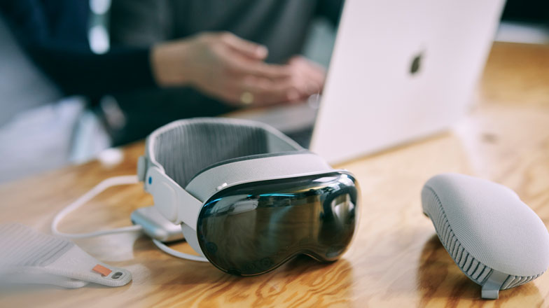 Auf einem Tisch liegt eine Apple Vision Pro, im Hintergrund sitzt jemand vor einem aufgeklappten Macbook.