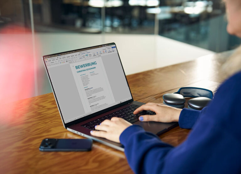 Eine Person sitzt an ihrem Laptop und schreibt eine Bewerbung, neben ihr liegen ein Smartphone und Overear Kopfhörer.