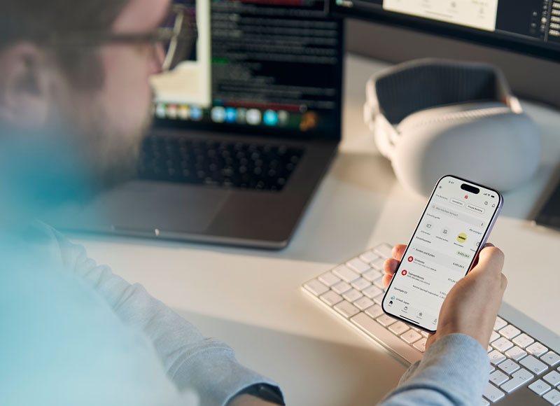 EIn Entwickler hält ein iPhone mit der Sparkassen App in der Hand und im Hintergrund siehnt man auf seinem Rechner Entwicklungscode.