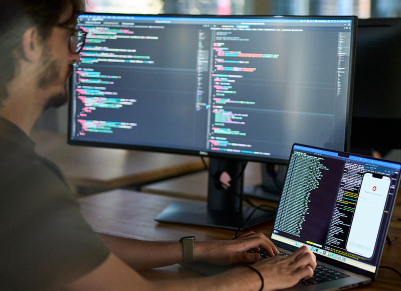 Entwickler sitzt an seibem Rechner, vor großen Monitoren und schreibt am Code der App Sparkasse.