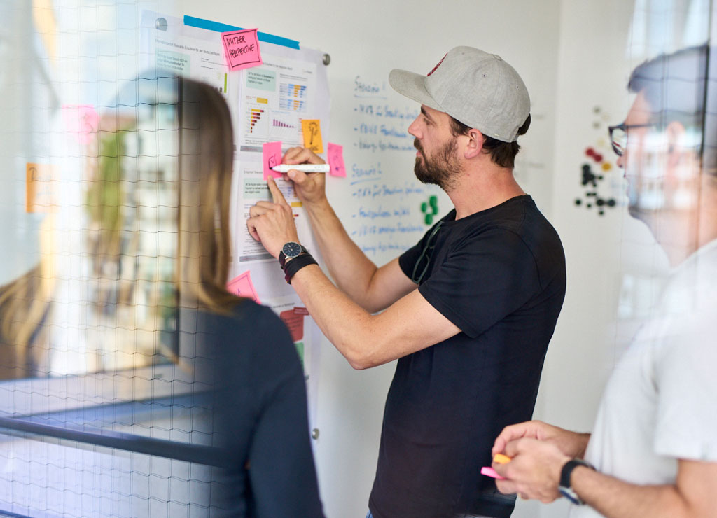 Drei Personen stehen vor einer Projektwand und beschreiben einen PostIt.