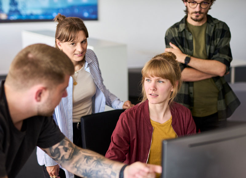 Vier junge Mitarbeitende stehn und sitzen gemeinsam vor einem Monitor und arbeiten gemeinsam an etwas.
