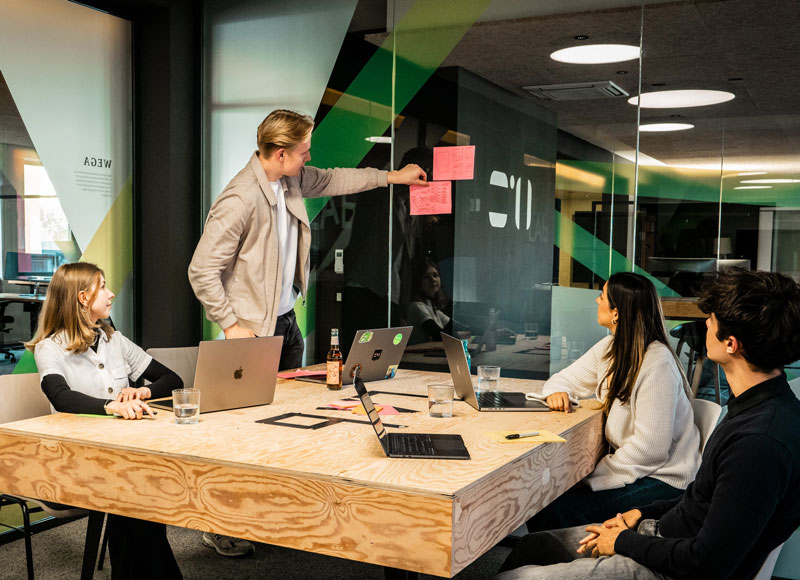 Vier duale Stundenten und Studentinnen sitzen mit Laptops, Notizbüchern und Getränken um einen Holztisch herum und arbeiten gemeinsam an einem Projekt in einem modernen, von Glaswänden umgebenen Büroraum.