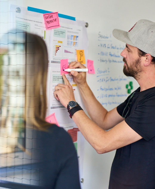 Drei Personen stehen vor einer Projektwand und beschreiben einen PostIt.