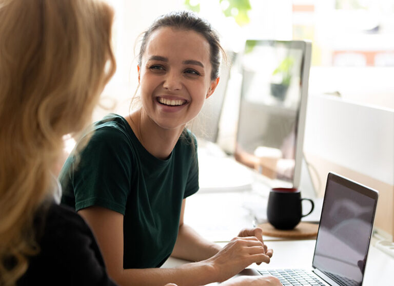 Zwei Frauen sitzen an einem Schreibtisch mit Computern; die eine lächelt und schaut die andere an.