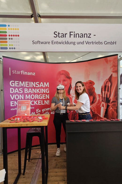 Eine Kollegin der Star Finanz steht mit VR-Brille auf gemeinsam mit einer anderen Kollegin auf dem Messestand einer Personalmesse.