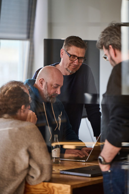 Fünf Männer stehen über ein Macbook geneigt und programmieren gemeinsam.