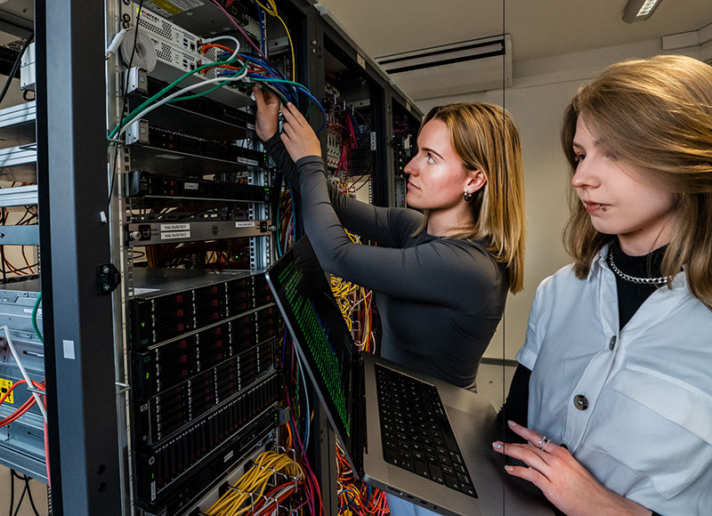 Zwei Auszubildende arbeiten in einem Serverraum; die eine schließt Kabel an ein Serverrack an, während die andere einen Laptop in der Hand hält und zusieht.