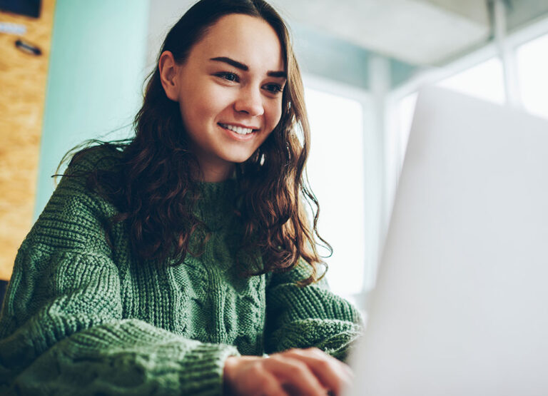 Eine junge Frau mit langen braunen Haaren, die einen grünen Pullover trägt, lächelt, während sie in einem hellen Raum mit großen Fenstern an einem Laptop arbeitet.