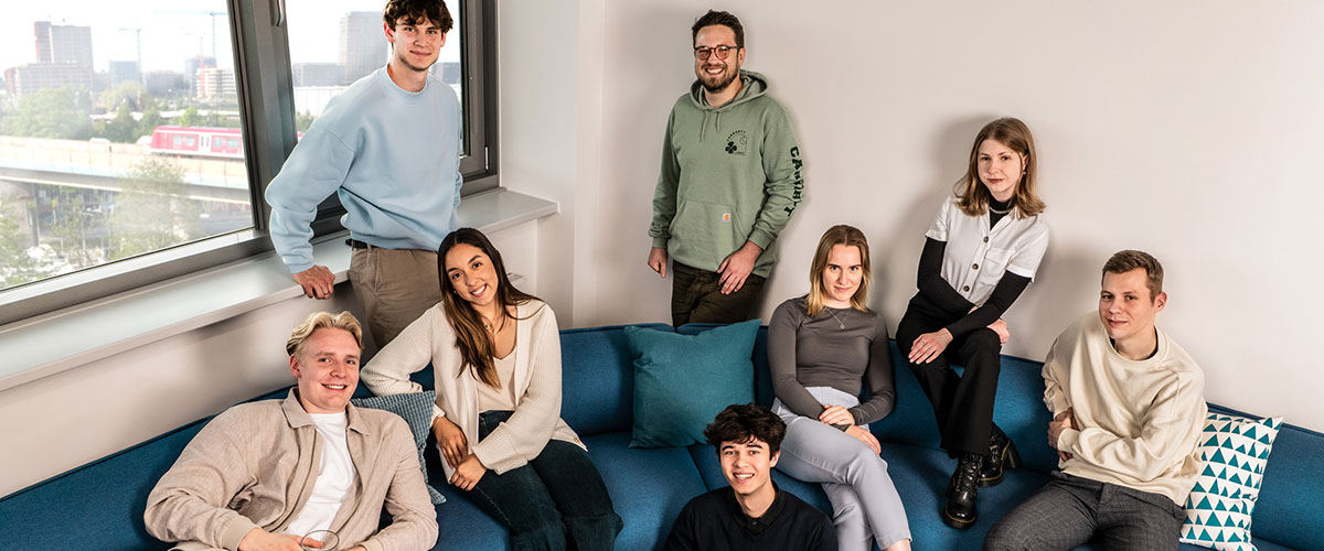 Acht Auszubildende und duale Student:innen sitzen und stehen um ein blaues Sofa herum in einem hellen, modernen Raum mit einem Fenster und Blick auf die Stadt.