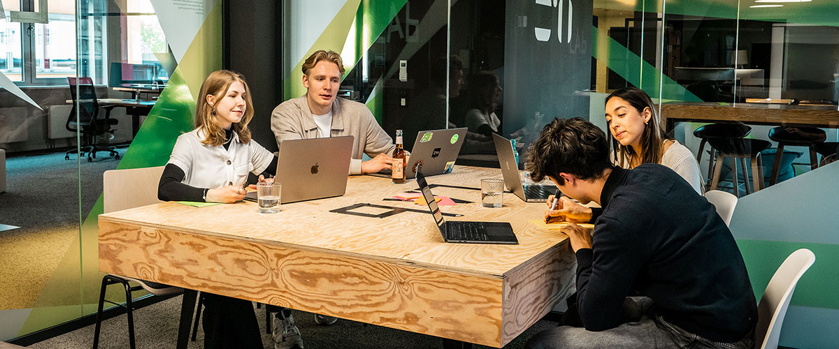 Vier duale Stundenten und Studentinnen sitzen mit Laptops, Notizbüchern und Getränken um einen Holztisch herum und arbeiten gemeinsam an einem Projekt in einem modernen, von Glaswänden umgebenen Büroraum.