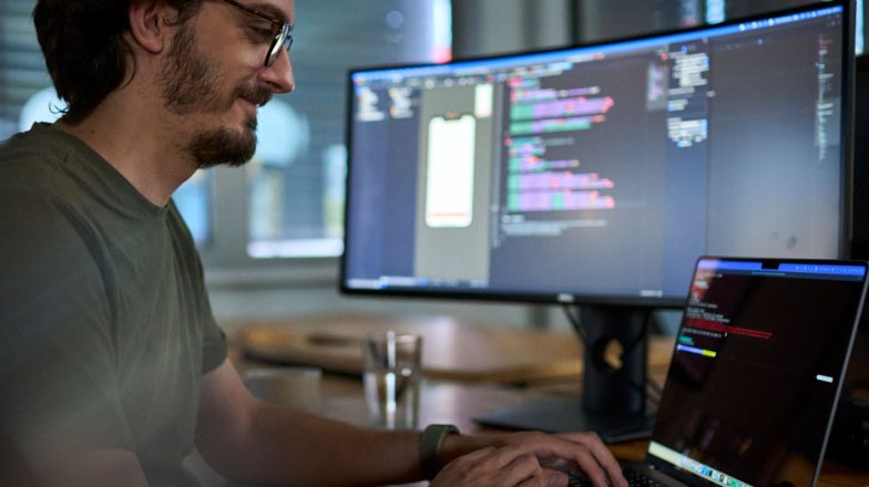 Ein Mitarbeiter der Star Finanz mit Brille sitzt lächelnd an einem Schreibtisch und arbeitet an einem Laptop. Auf dem großen Monitor vor ihm ist farbiger Programmcode sowie ein Smartphone-Layout zu sehen.