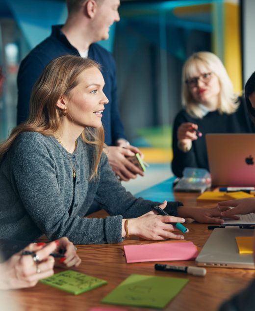 Mehrere Mitarbeitende des Sparkassen Innovation Hubs arbeiten gemeinsam an einem Tisch mit Laptops, Stiften und bunten Notizzetteln. Eine Frau in grauer Strickjacke spricht engagiert, während andere zuhören und mitschreiben. Kreative Teamatmosphäre in einem modernen Büro.