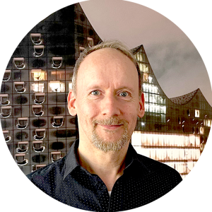 Porträtfoto von Ingo Helmke, ein Mannes mit kurzem Haar und hellem Bart, der ein dunkles Hemd trägt. Im Hintergrund ist die beleuchtete Glasfassade eines modernen Gebäudes mit markanter Architektur zu sehen.