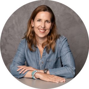Portrait von Dana Gielnik, Leiterin People, Culture & Places, die lächelnd am Tisch sitzt, vor grauem Hintergrund.