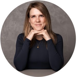 Portrait von Dana Paul, People-Spezialistin, die lächelnd mit den Händen unter dem Kinn am Tisch sitzt, vor grauem Hintergrund.