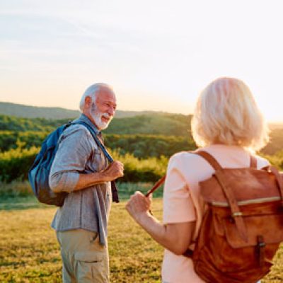 Zwei ältere Menschen mit Rucksäcken lächeln sich an, während sie bei Sonnenuntergang durch eine Wiese wandern
