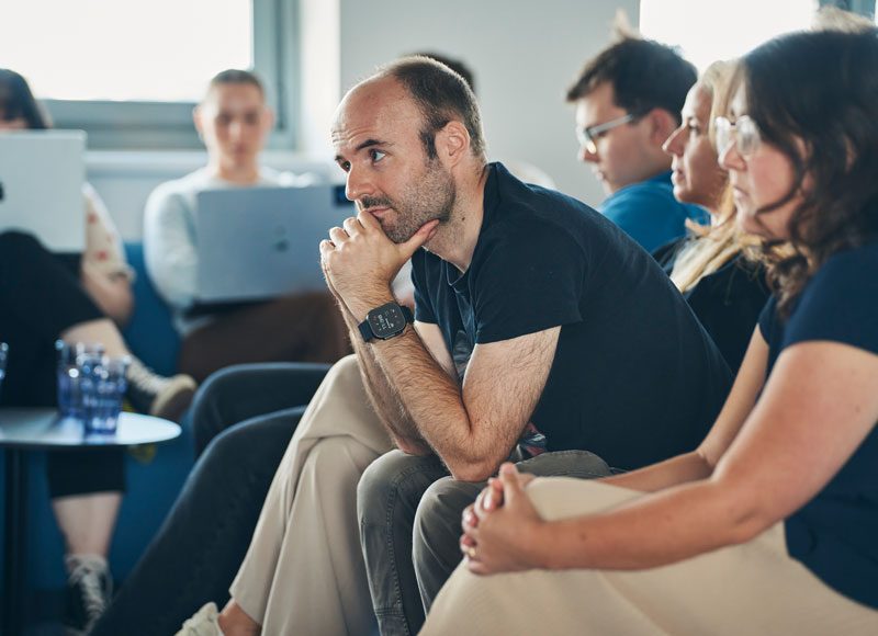 Ein Mann sitzt im Vordergrund vorbeugt in einer Gruppe von Kolleg:innen und hört aufmerksam einem Vortrag zu.