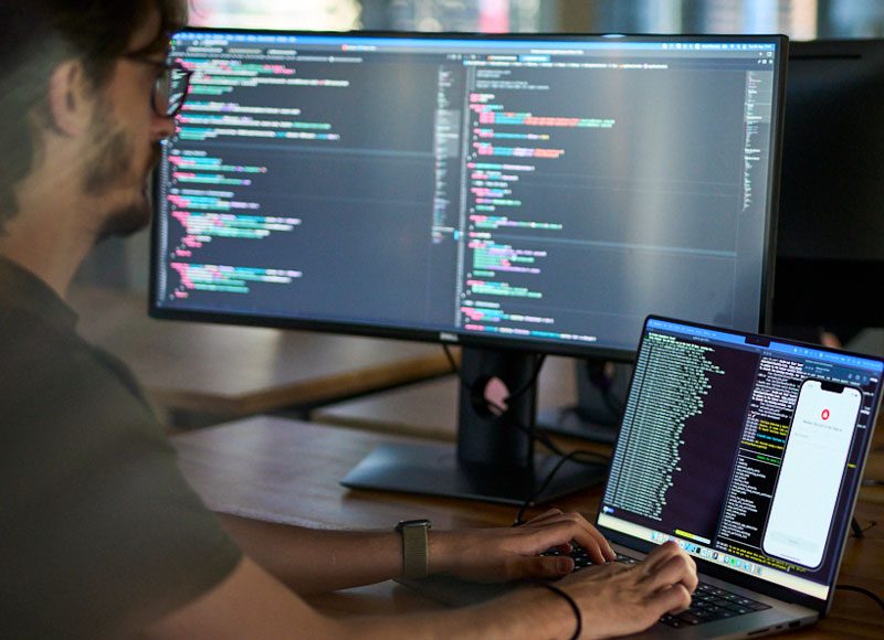 Entwickler sitzt an seibem Rechner, vor großen Monitoren und schreibt am Code der App Sparkasse.