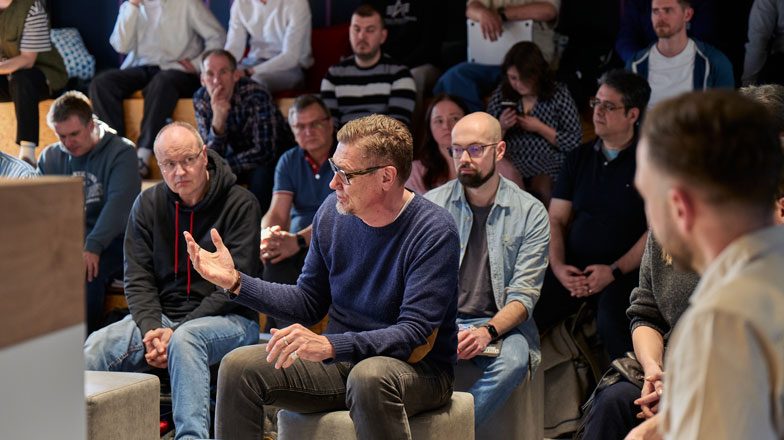 Ein Mann sitzt im Vordergrund im Rahmen eines größeren Meetings und um ihn herum zig andere Personen, die ihm zuhören.