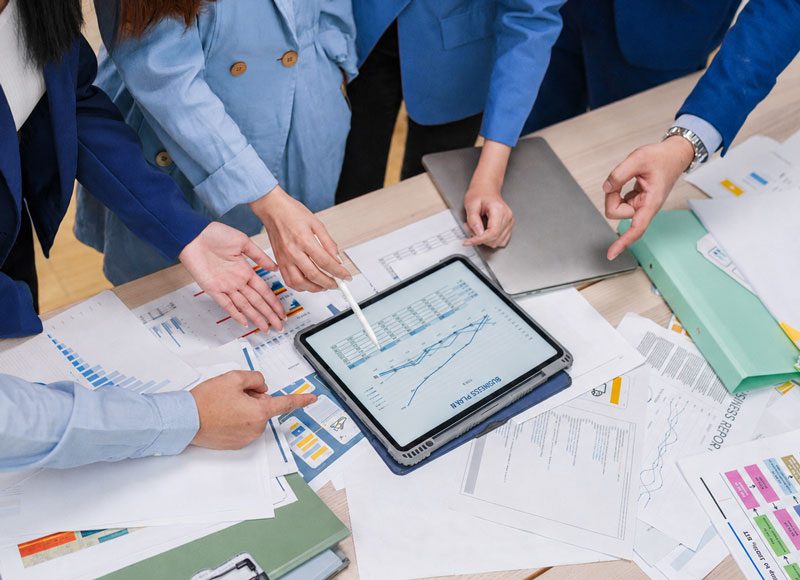 Vier Personen stehen gemeinsam um einen Tisch herum, auf dem Planungs- und Strategiepapiere liegen und diskutieren die Planzahlen auf dem Tablet.