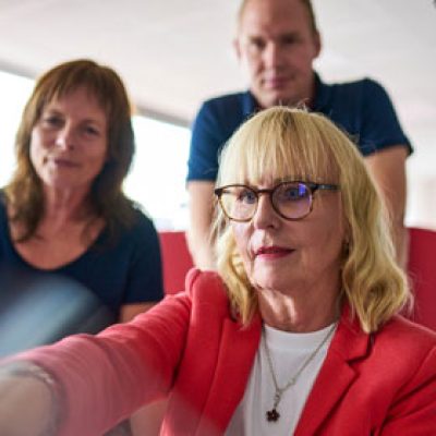 Eine ältere Frau schaut auf einen Bildschirm und zwei weitere Personen stehen hinter ihr und schauen ihr über die Schulter