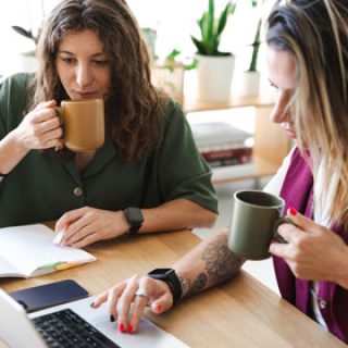 Zwei Frauen sitzen gemeinsam vor einem Laptop und trinken dabei etwas aus Tassen, die sie in der Hand halten.