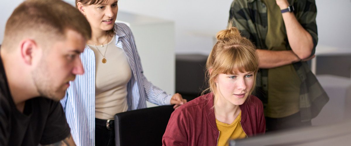 Vier junge Mitarbeitende stehn und sitzen gemeinsam vor einem Monitor und arbeiten gemeinsam an etwas.