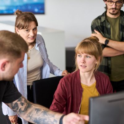 Vier junge Mitarbeitende stehn und sitzen gemeinsam vor einem Monitor und arbeiten gemeinsam an etwas.