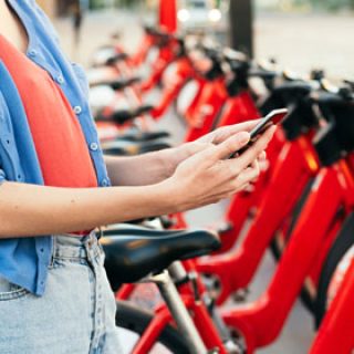 Eine Person nutzt ihr Smartphone, während sie neben einer Reihe roter Leihfahrräder steht, die auf einer Straße in der Stadt geparkt sind.
