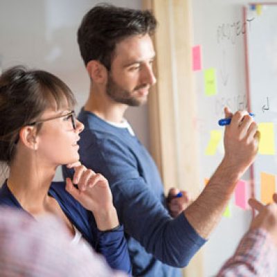 Drei Personen stehen vor einer Projektwand und ein Mann schreibt etwas an die Wand.