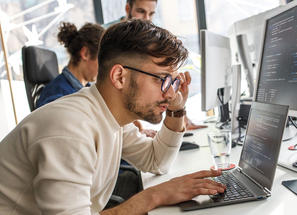 Im Vordergrund sitzt ein Entwickler vor seinem Laptop und prüft konzentriert den Code.