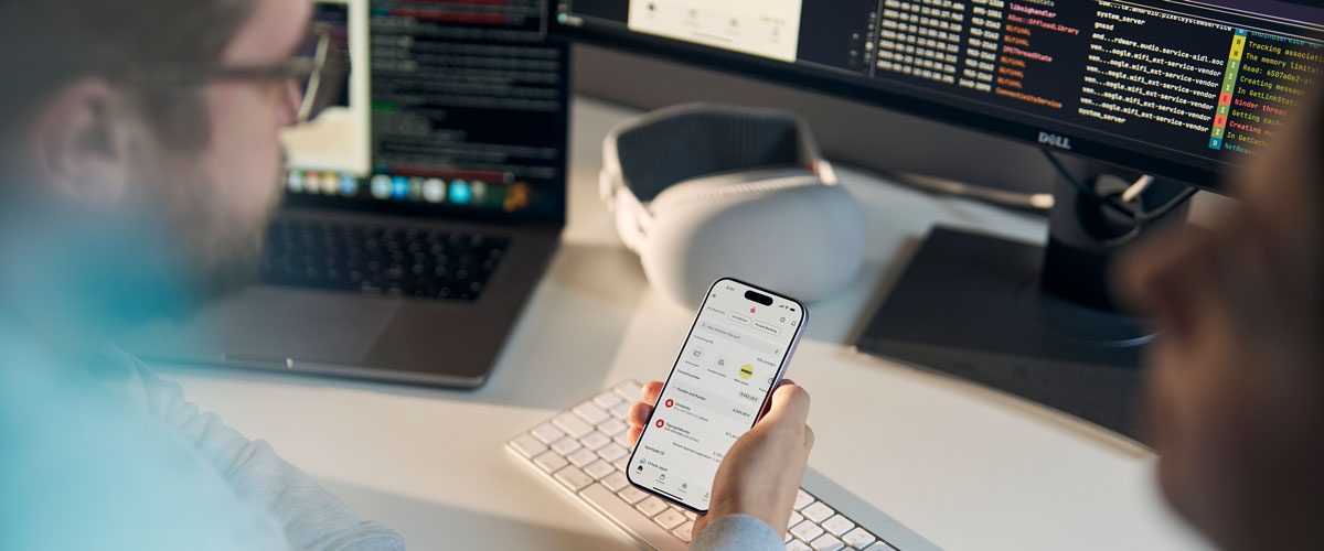 EIn Entwickler hält ein iPhone mit der Sparkassen App in der Hand und im Hintergrund siehnt man auf seinem Rechner Entwicklungscode.