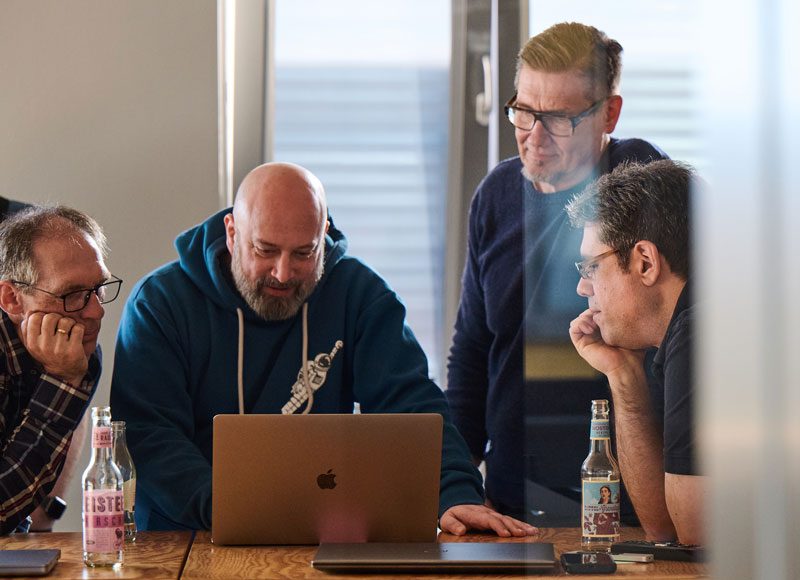 Vier Männer stehen über ein Macbook geneigt und programmieren gemeinsam.