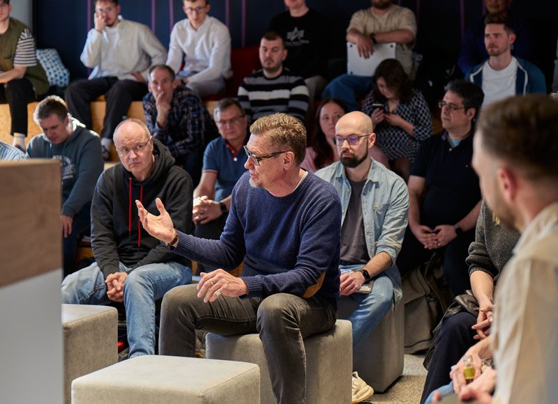 Im Rahmen eines Workshops sitzt eine große Gruppe zusammen und hört einem in der Mitte sitzenden Mann bei seinem Beitrag zu.